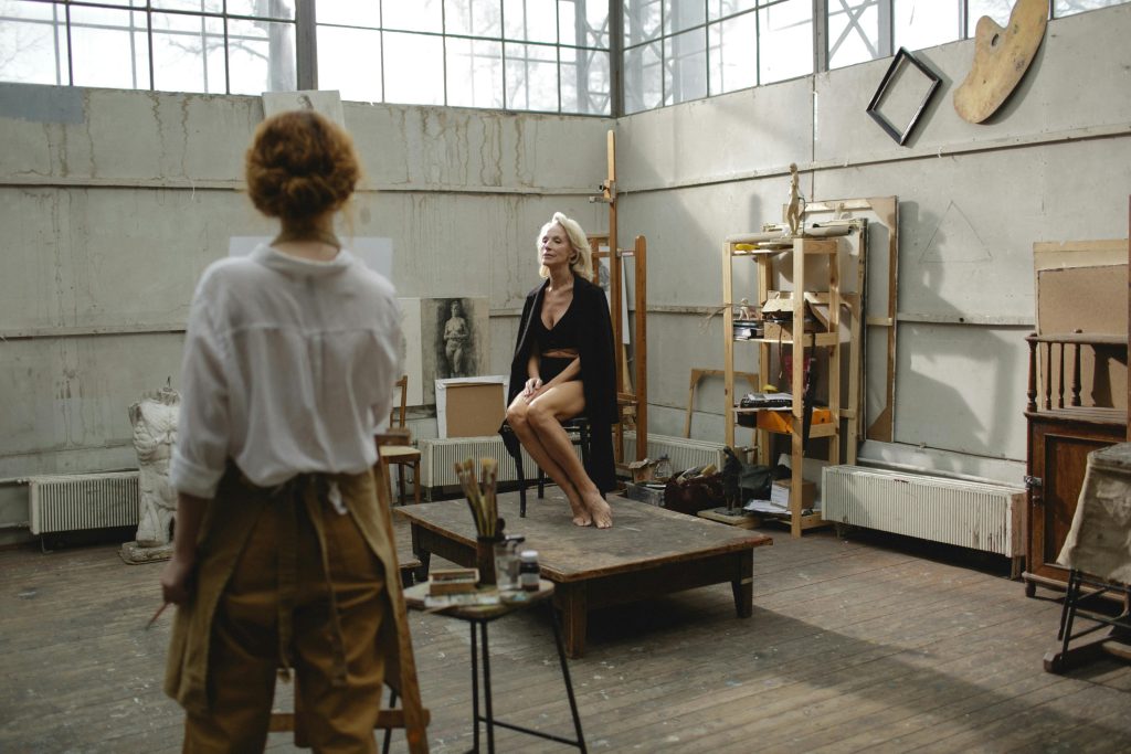 An elderly artist paints a senior model posing in a bright art studio, focusing on creativity and concentration.